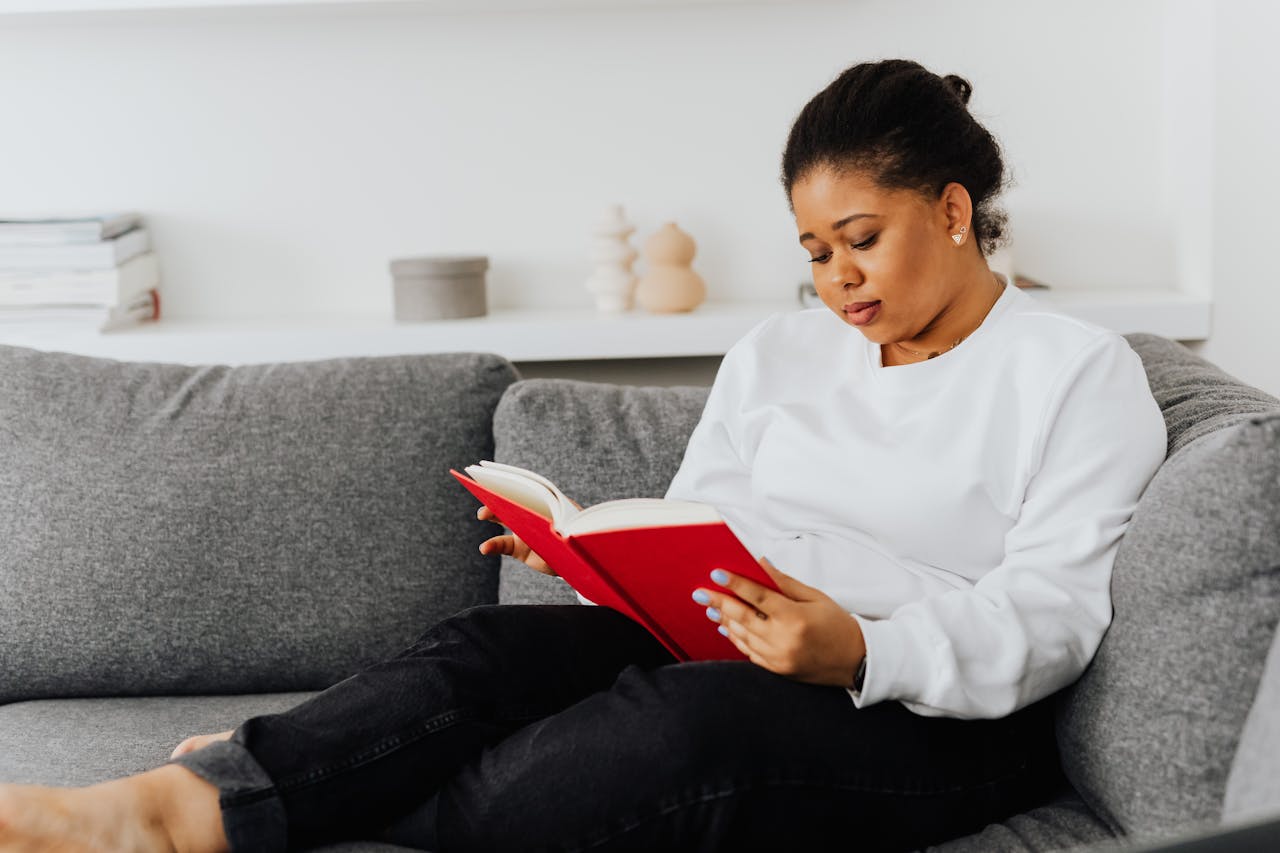 African American woman wearing casual attire reading a book on a comfortable sofa indoors.